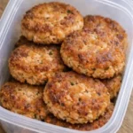 Four golden-crusted salmon patties in a cast iron skillet, crispy at the edges, served with lemon wedges and a small bowl of creamy dipping sauce on a white linen surface.