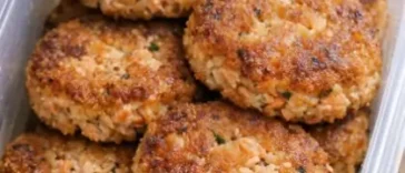 Four golden-crusted salmon patties in a cast iron skillet, crispy at the edges, served with lemon wedges and a small bowl of creamy dipping sauce on a white linen surface.
