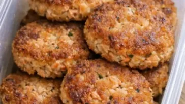 Four golden-crusted salmon patties in a cast iron skillet, crispy at the edges, served with lemon wedges and a small bowl of creamy dipping sauce on a white linen surface.