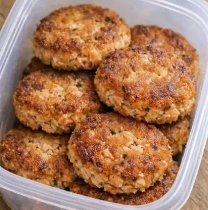 Four golden-crusted salmon patties in a cast iron skillet, crispy at the edges, served with lemon wedges and a small bowl of creamy dipping sauce on a white linen surface.