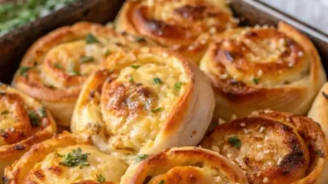A close-up overhead shot of small garlic rolls made from rolled sandwich crusts, arranged in a round cast iron skillet, brushed with glistening garlic herb butter and sprinkled with coarse salt and chopped parsley, resting on a dark wooden table.