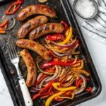 A cast iron skillet filled with sliced Italian sausage, colorful bell pepper strips in red, yellow, and orange, and caramelized onion ribbons glistening in a light pan sauce, shot from above on a dark wooden surface.