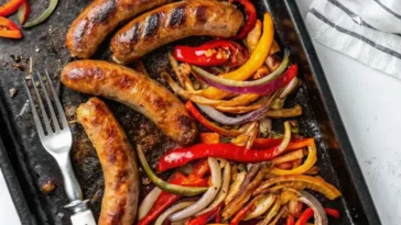 A cast iron skillet filled with sliced Italian sausage, colorful bell pepper strips in red, yellow, and orange, and caramelized onion ribbons glistening in a light pan sauce, shot from above on a dark wooden surface.