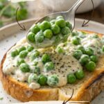 A wooden spoon stirring bright green frozen peas into a bubbling cream sauce in a small saucepan on a gas stovetop.