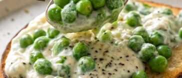 A wooden spoon stirring bright green frozen peas into a bubbling cream sauce in a small saucepan on a gas stovetop.