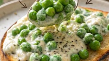 A wooden spoon stirring bright green frozen peas into a bubbling cream sauce in a small saucepan on a gas stovetop.