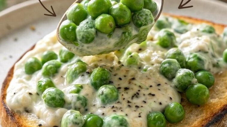 A wooden spoon stirring bright green frozen peas into a bubbling cream sauce in a small saucepan on a gas stovetop.