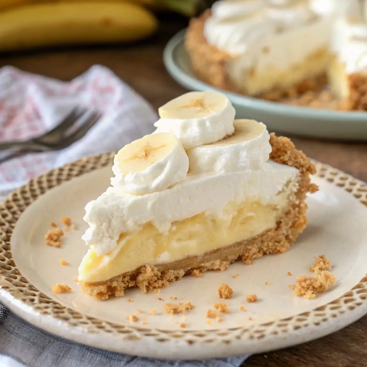 A single slice of no-bake banana cream pie on a white ceramic plate, showing distinct layers: a golden graham cracker crust at the bottom, a thick cream cheese and pudding filling with banana slices, and a tall swirl of whipped cream on top, photographed in natural window light.


