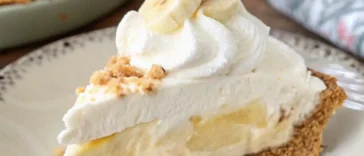 A chilled no-bake banana cream pie in a glass dish, topped with swirls of fresh whipped cream and overlapping banana slices, shot from above on a linen-covered wooden table with a vintage fork beside it.