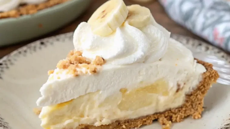 A chilled no-bake banana cream pie in a glass dish, topped with swirls of fresh whipped cream and overlapping banana slices, shot from above on a linen-covered wooden table with a vintage fork beside it.