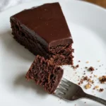 Overhead shot of a round kladdkaka dusted with powdered sugar on a white ceramic plate, one slice removed to show the dark fudgy center, in warm natural light on a linen surface.