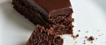 Overhead shot of a round kladdkaka dusted with powdered sugar on a white ceramic plate, one slice removed to show the dark fudgy center, in warm natural light on a linen surface.