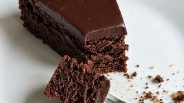Overhead shot of a round kladdkaka dusted with powdered sugar on a white ceramic plate, one slice removed to show the dark fudgy center, in warm natural light on a linen surface.