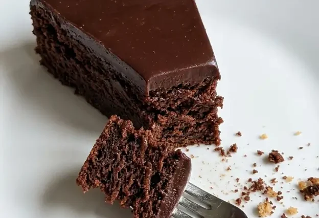 Overhead shot of a round kladdkaka dusted with powdered sugar on a white ceramic plate, one slice removed to show the dark fudgy center, in warm natural light on a linen surface.