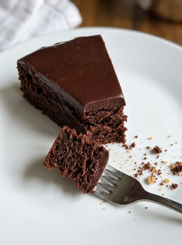 Overhead shot of a round kladdkaka dusted with powdered sugar on a white ceramic plate, one slice removed to show the dark fudgy center, in warm natural light on a linen surface.
