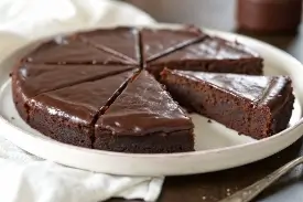 Overhead shot of a round kladdkaka dusted with powdered sugar on a white ceramic plate, one slice removed to show the dark fudgy center, in warm natural light on a linen surface.