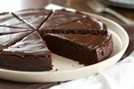 Overhead shot of a round kladdkaka dusted with powdered sugar on a white ceramic plate, one slice removed to show the dark fudgy center, in warm natural light on a linen surface.